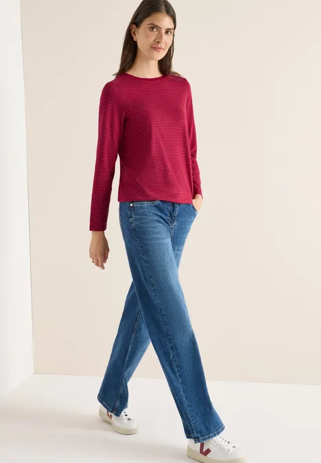 Frau in rotem Langarmshirt, blauer Jeans und weißen Sneakern vor hellem Hintergrund.